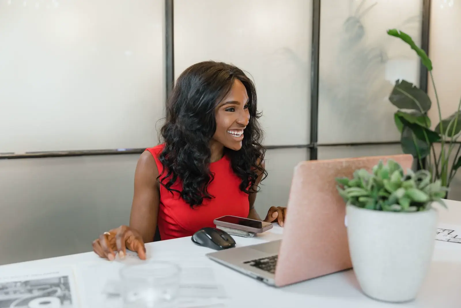 A woman smiling at a desk with a laptop and phone, surrounded by plants.